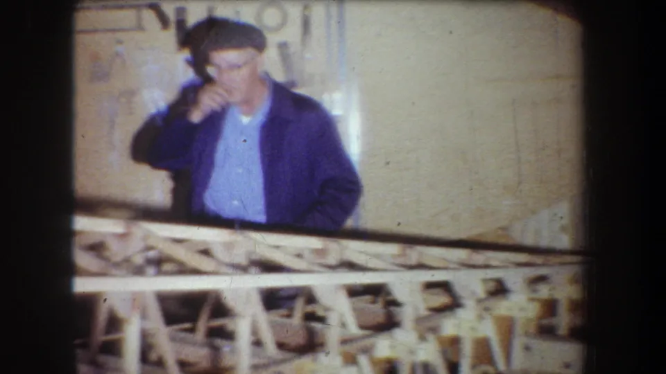 1965 Yucaipa home-movie frame showing the man in a blue jacket behind the wooden build, with his hand lifted near his face.