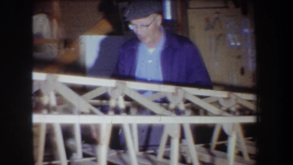 1965 Yucaipa frame showing a man in a flat cap looking down across an intricate unfinished wooden structure in a private workshop.