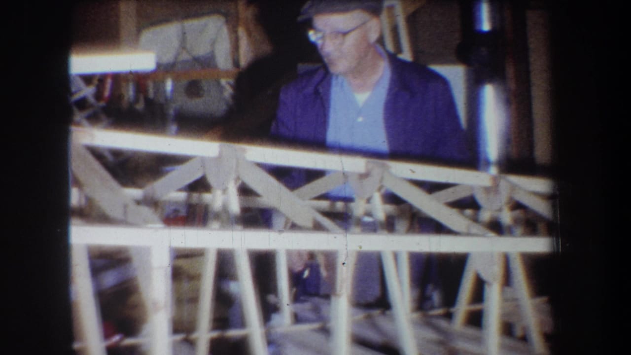 1965 Yucaipa home-movie frame showing a man in a cap and glasses working near a wooden sailplane frame inside a garage workshop.