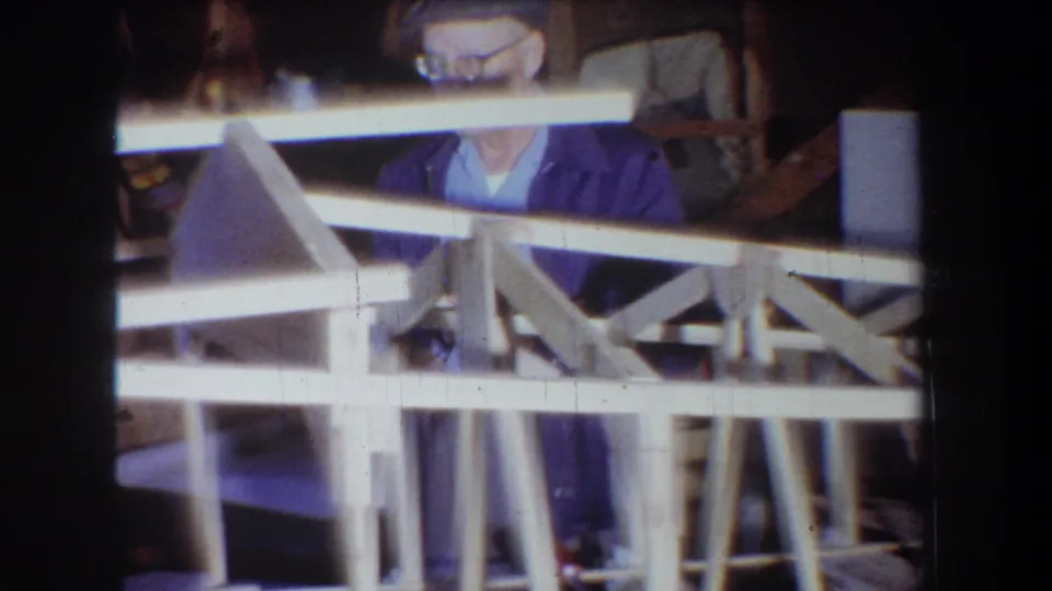 1965 Yucaipa footage shows the man partly obscured by a pale wooden sailplane frame and surrounding garage workshop materials.