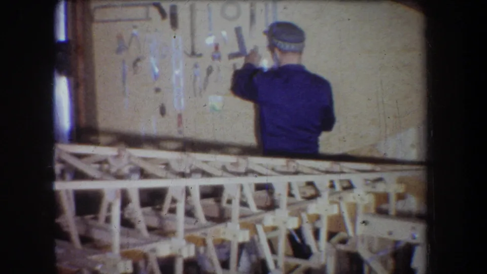 1965 Yucaipa home-movie view from behind as the man reaches toward wall-mounted tools above the wooden sailplane frame.
