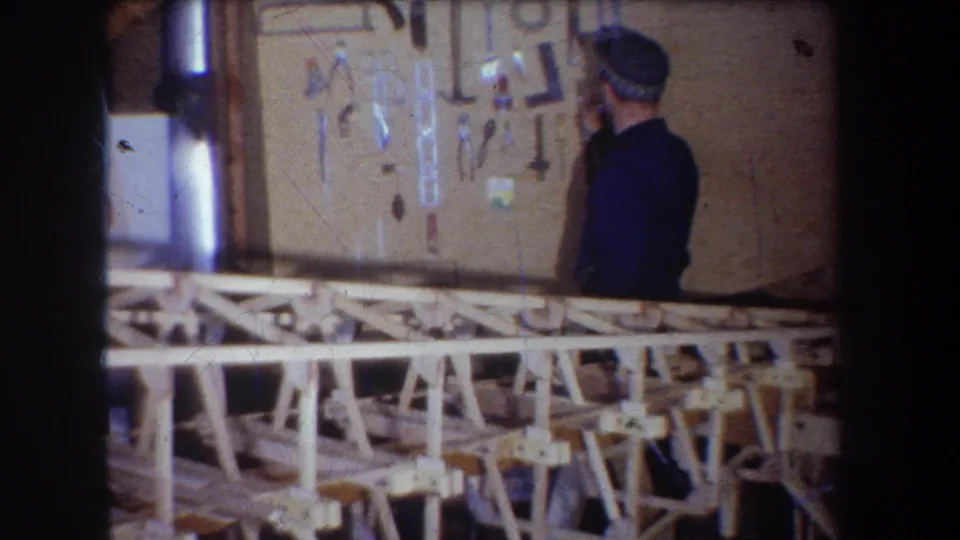 In a 1965 Yucaipa-area workshop scene, a man in a blue jacket and cap faces a wall of hanging tools behind pale wooden benches.