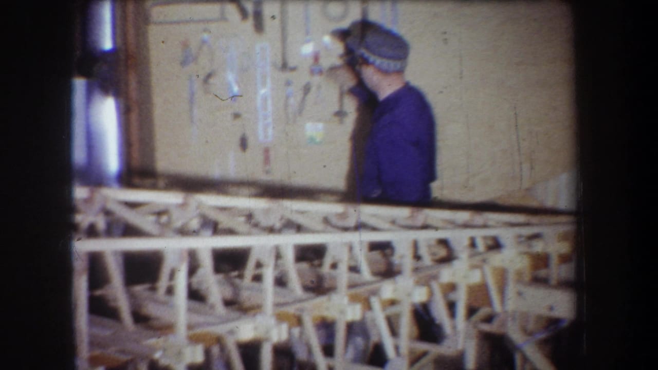 The man turns toward the tool display in the 1965 home-movie workshop, with workbench framing across the foreground.