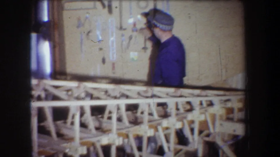 The man turns toward the tool display in the 1965 home-movie workshop, with workbench framing across the foreground.