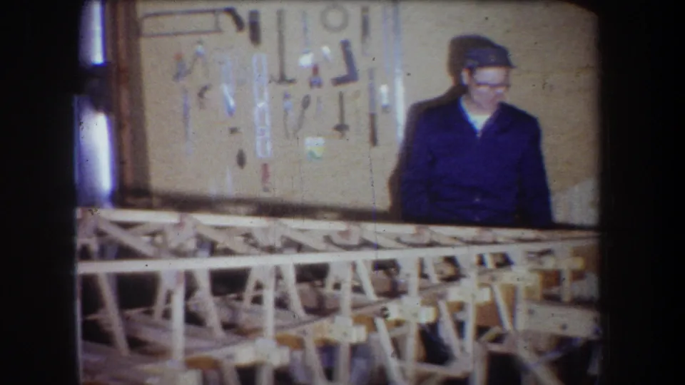 The man stands closer to the center of the workshop frame, facing forward near the hanging tools and bench structure.