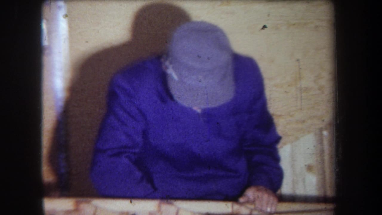 1965 home-movie frame from Yucaipa area showing an adult man in a purple sweater and gray cap leaning over a wooden surface in a paneled room.