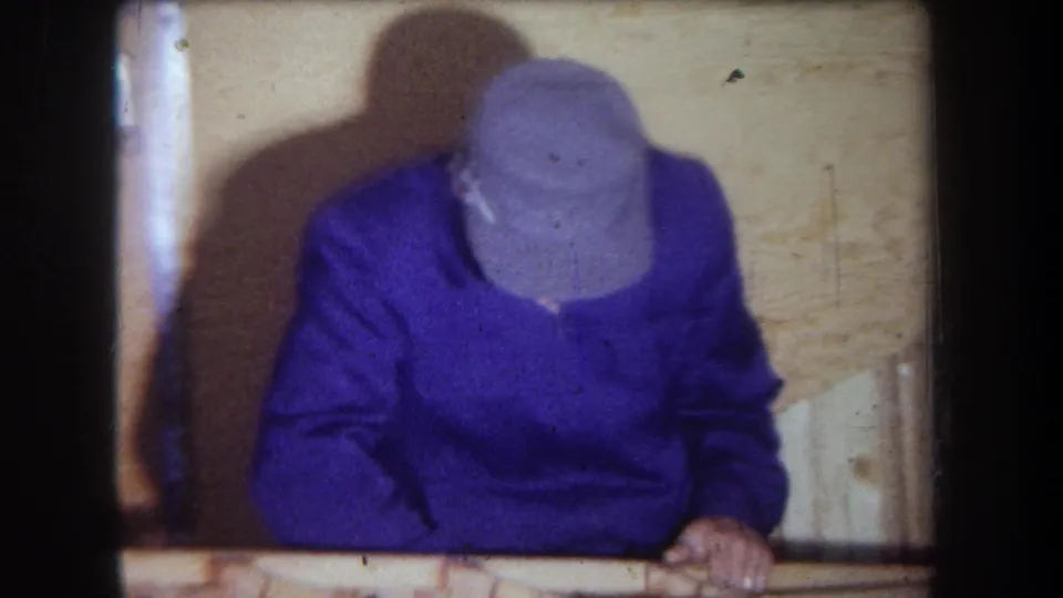 1965 home-movie frame from Yucaipa area showing an adult man in a purple sweater and gray cap leaning over a wooden surface in a paneled room.