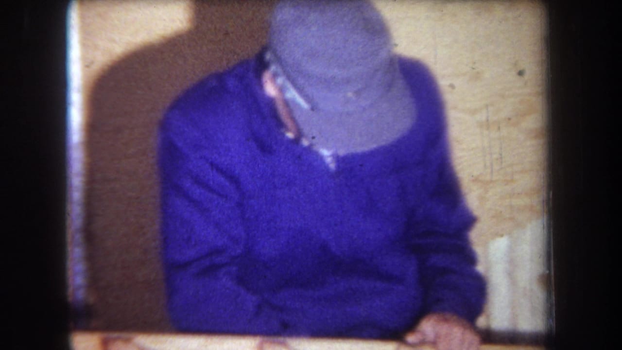 Adult man in a cap and purple sweater bends over a wooden surface in a dim interior, consistent with the 1965 home-movie clip metadata.