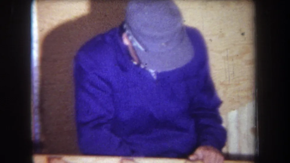 Adult man in a cap and purple sweater bends over a wooden surface in a dim interior, consistent with the 1965 home-movie clip metadata.