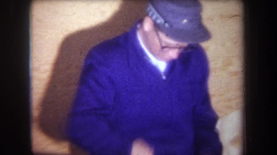 Adult man in a purple sweater and gray cap turns beside the wooden surface, with motion blur and 8mm grain visible in the interior frame.