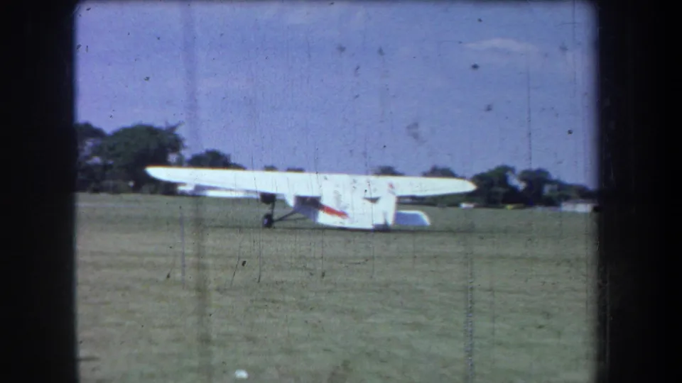 White single-engine propeller airplane with a red stripe sits on a grassy airfield with trees in the background, from the 1965 home-movie clip.