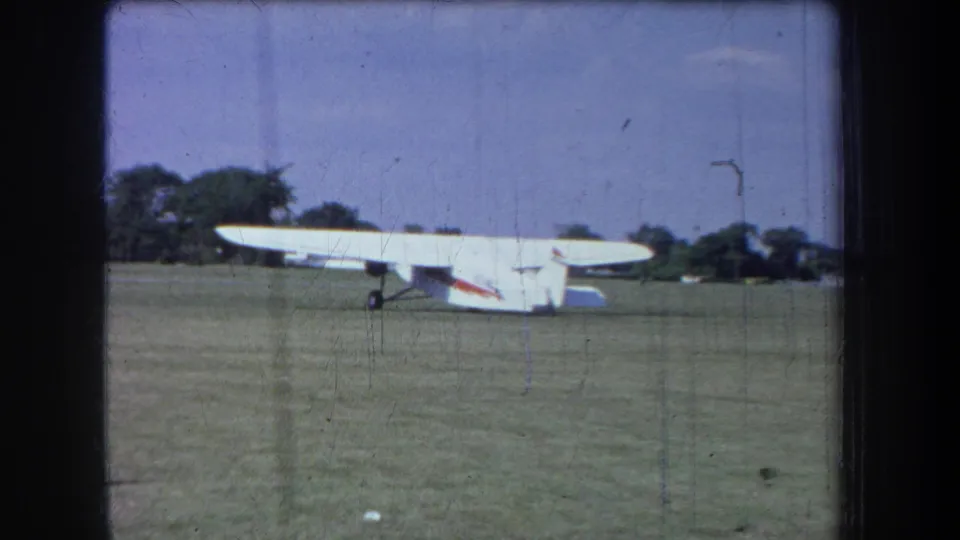 1965 Yucaipa-area 8mm frame showing a white light airplane on a grass airfield with trees in the background.