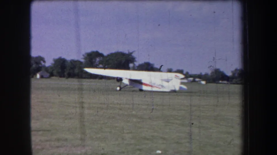 1965 8mm frame of a small white airplane on a rural California grass field with visible film wear.