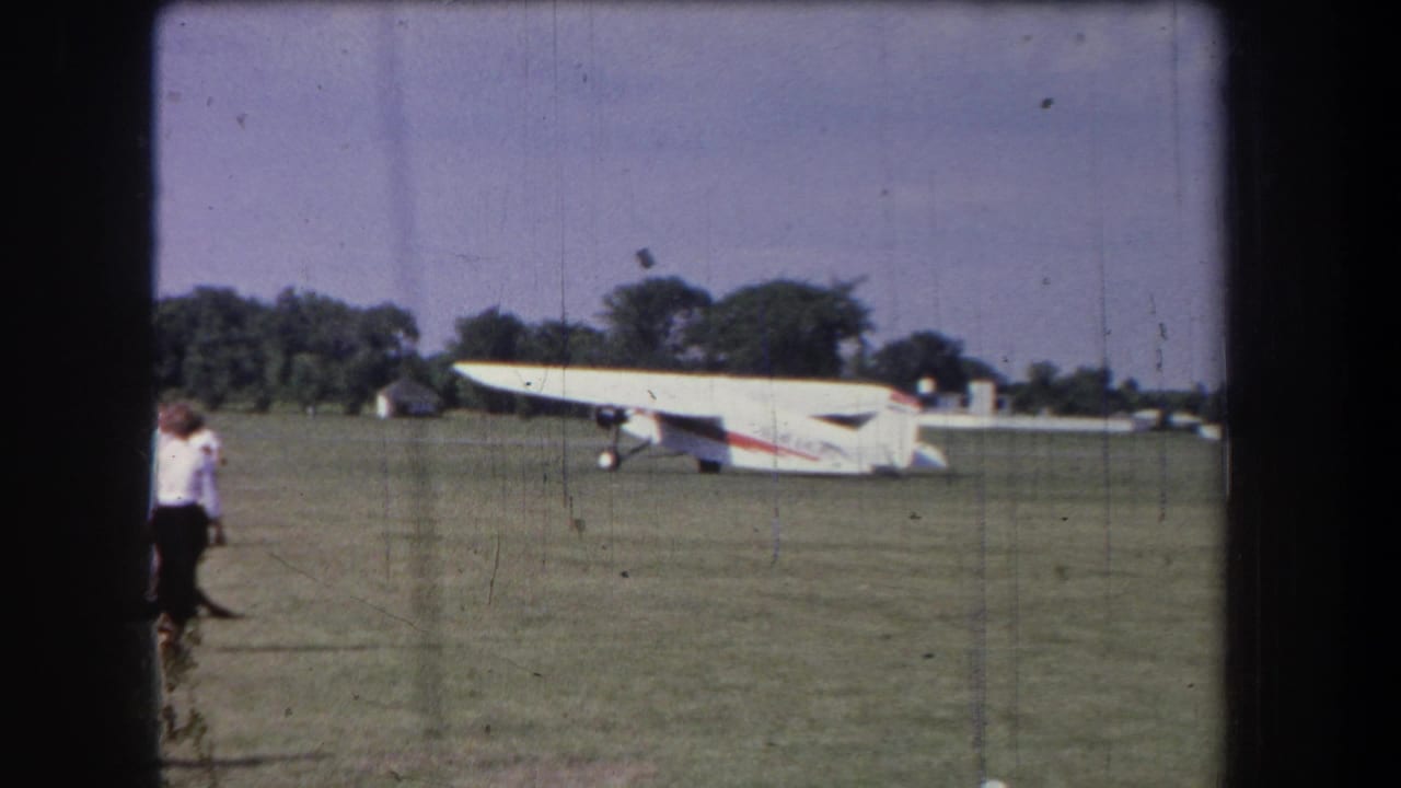 1965 Yucaipa-area 8mm frame showing a white airplane with red striping on grass as spectators stand near the field edge.