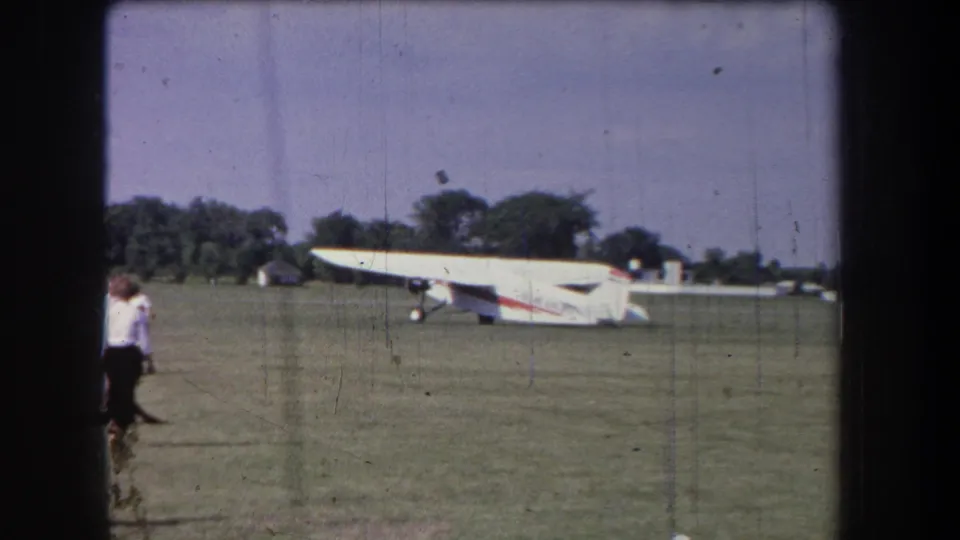 1965 Yucaipa-area 8mm frame showing a white airplane with red striping on grass as spectators stand near the field edge.
