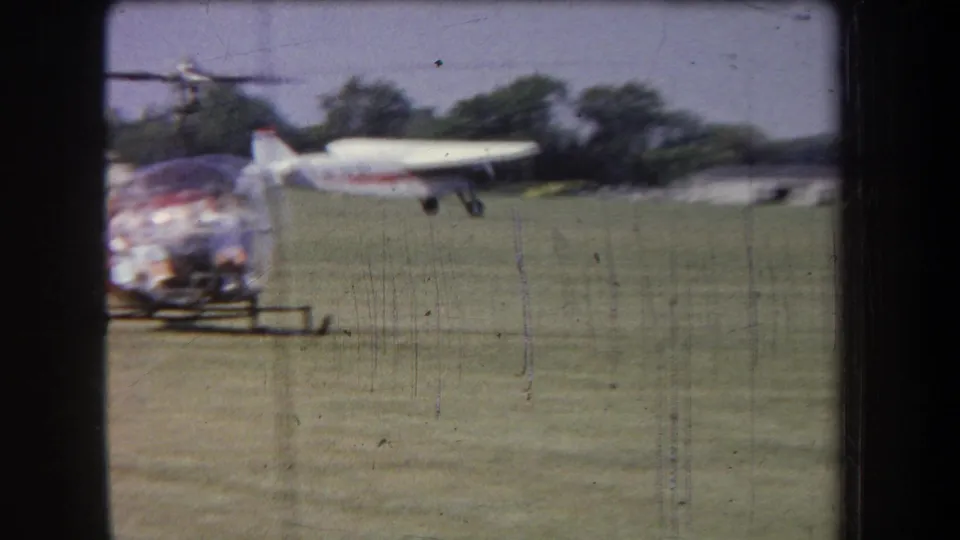 A bubble-canopy helicopter sits in the foreground while a small white airplane moves across the grassy Yucaipa-area airfield.