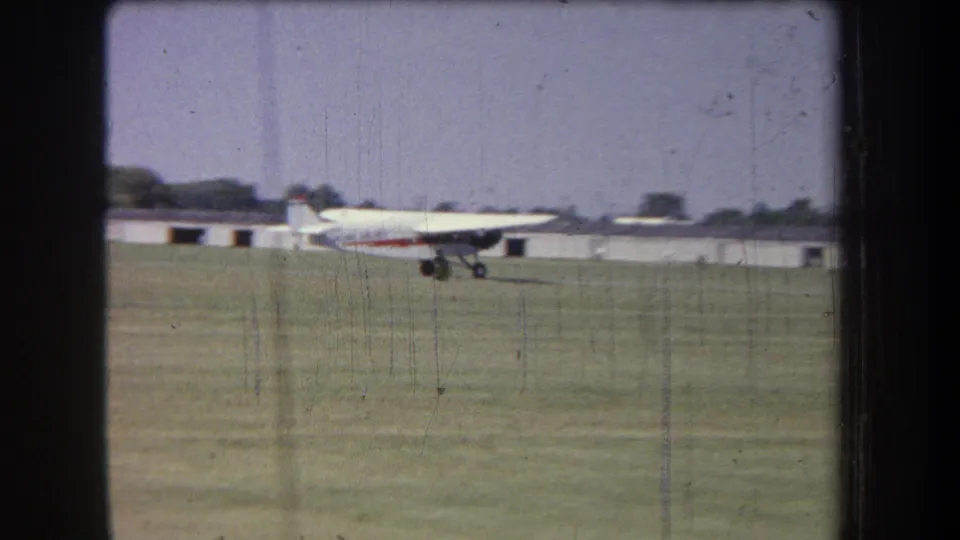 The small airplane appears to lift off from the grassy airfield, with low hangar-like buildings and trees behind it.