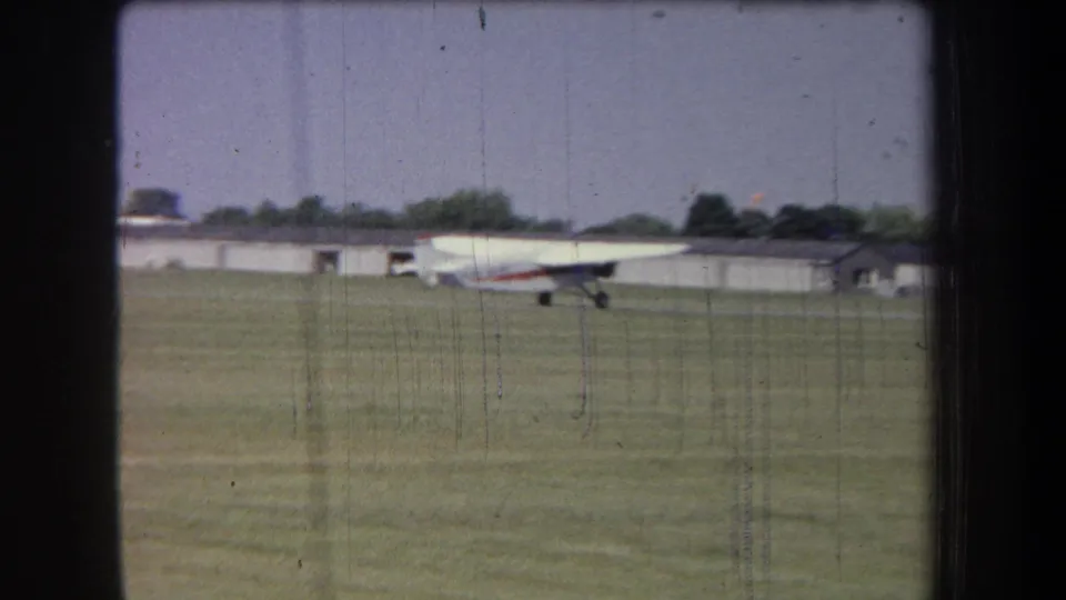 The white airplane continues across the grass in the 1965 Yucaipa-area scene, framed against low buildings and a tree line.