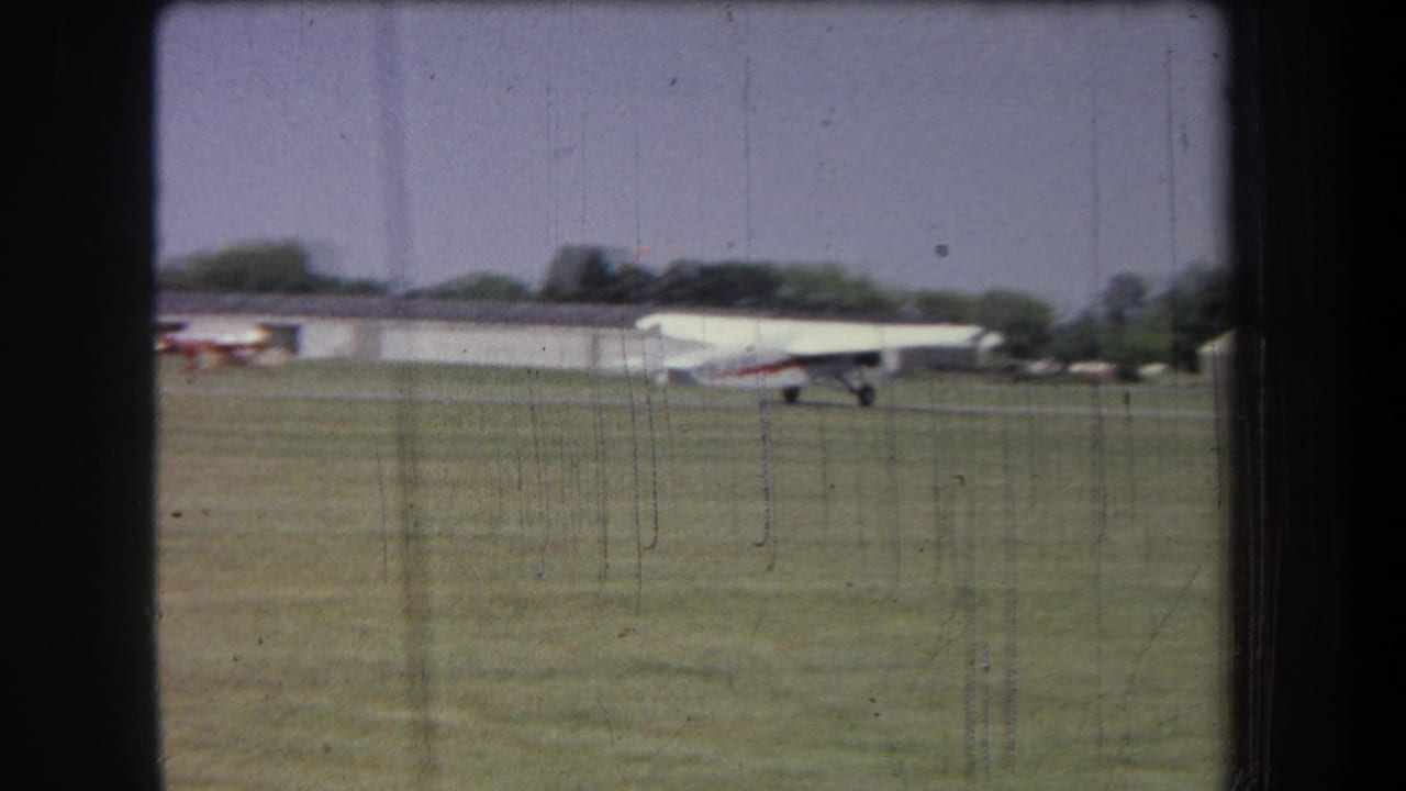 1965 8mm frame from Volmer Yucaipa, California, showing a small white plane with red accents on a grass airfield near hangars.