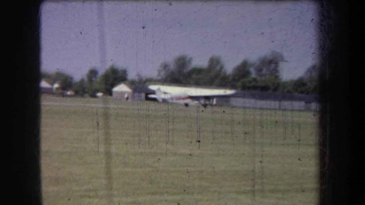 Home-movie frame showing a small white aircraft with red trim on a grassy Yucaipa airfield under a pale sky.