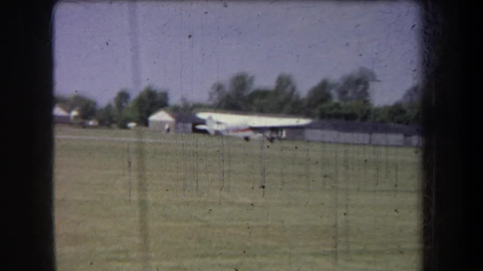 Home-movie frame showing a small white aircraft with red trim on a grassy Yucaipa airfield under a pale sky.
