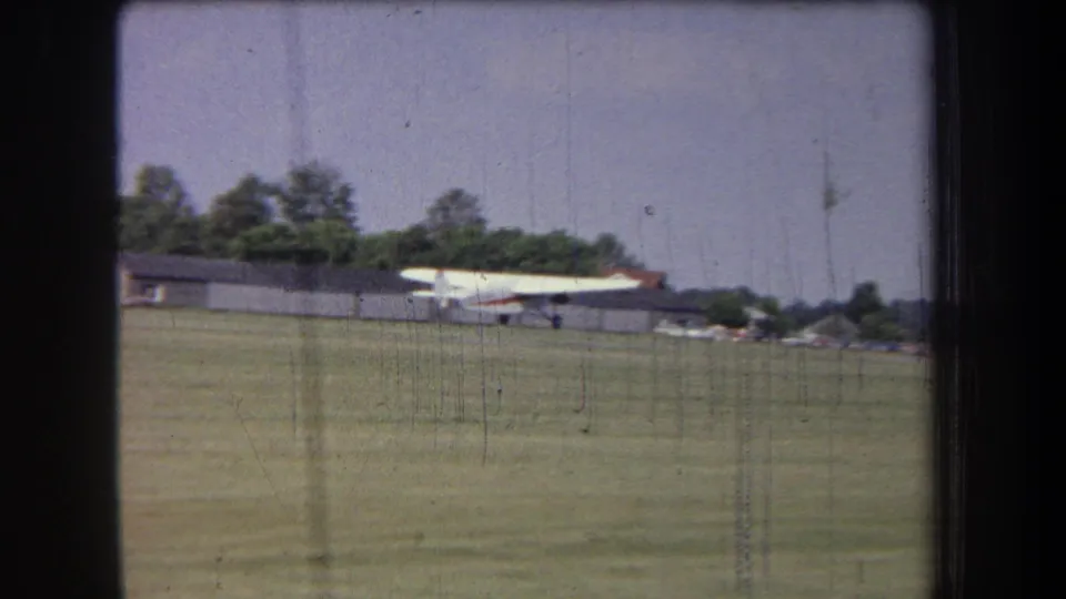 1965 8mm view of a small white plane moving across a grass airfield in Volmer Yucaipa, with low buildings behind it.