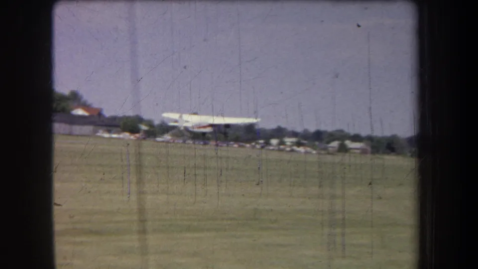 Final frame of the 1965 Yucaipa airfield scene, with the small white plane on grass and hangars visible beyond it.