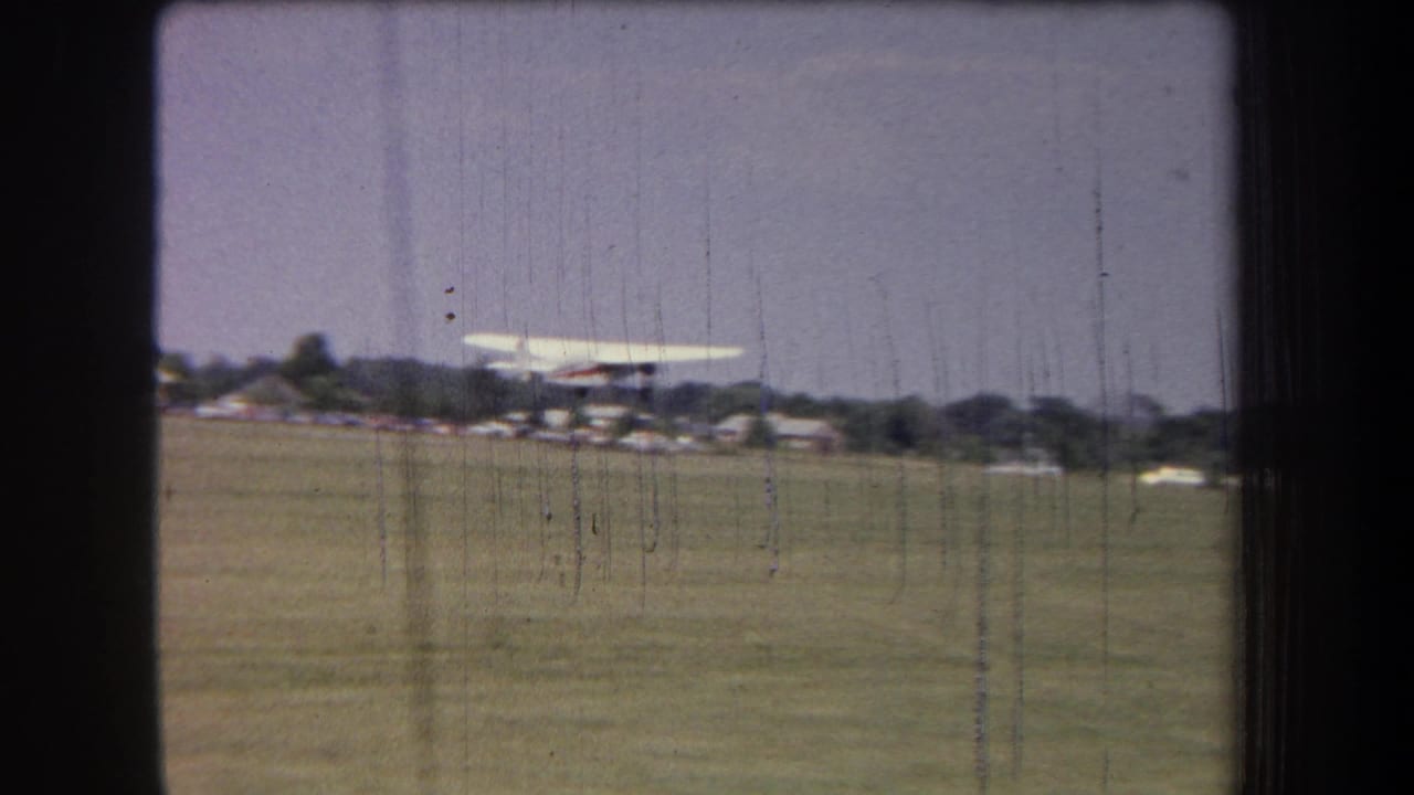 1965 8mm frame from Volmer Yucaipa, California, showing a small propeller plane just above a grass airfield.