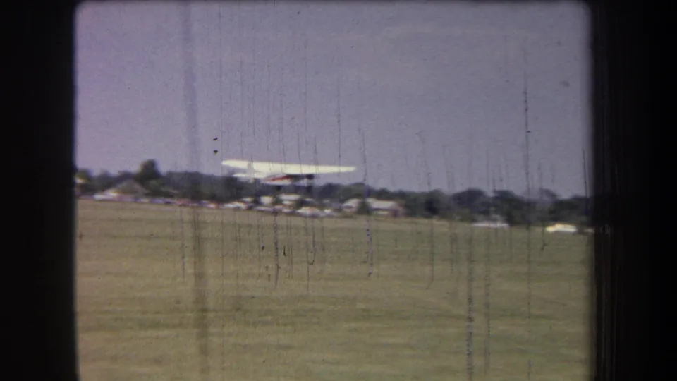 1965 8mm frame from Volmer Yucaipa, California, showing a small propeller plane just above a grass airfield.