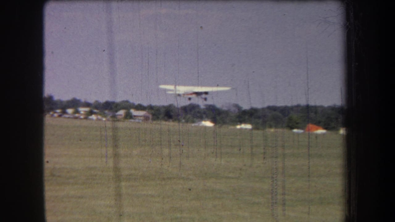 Small aircraft airborne over an open grass runway with trees and low buildings beyond.