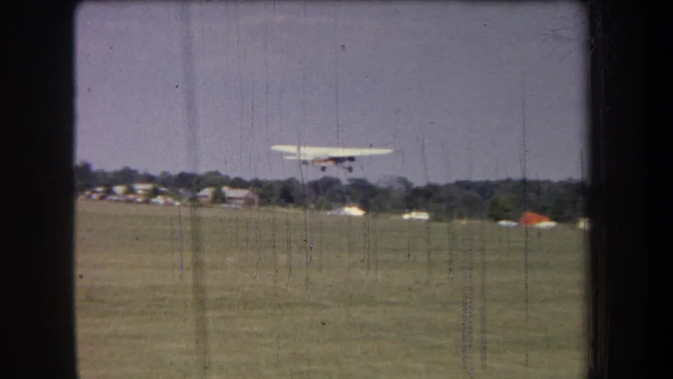 Small propeller aircraft climbs over a rural grass airfield in 1965 Volmer Yucaipa home-movie footage.