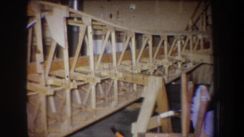 Final frame shows the braced wooden assembly continuing across the scene, with workshop materials partly visible near the right edge.