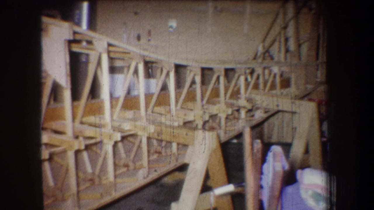 1965 home-movie frame from Yucaipa showing a pale wooden structure under construction in a workshop, with trusses, posts, and a partly visible person.