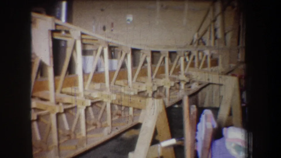 1965 home-movie frame from Yucaipa showing a pale wooden structure under construction in a workshop, with trusses, posts, and a partly visible person.