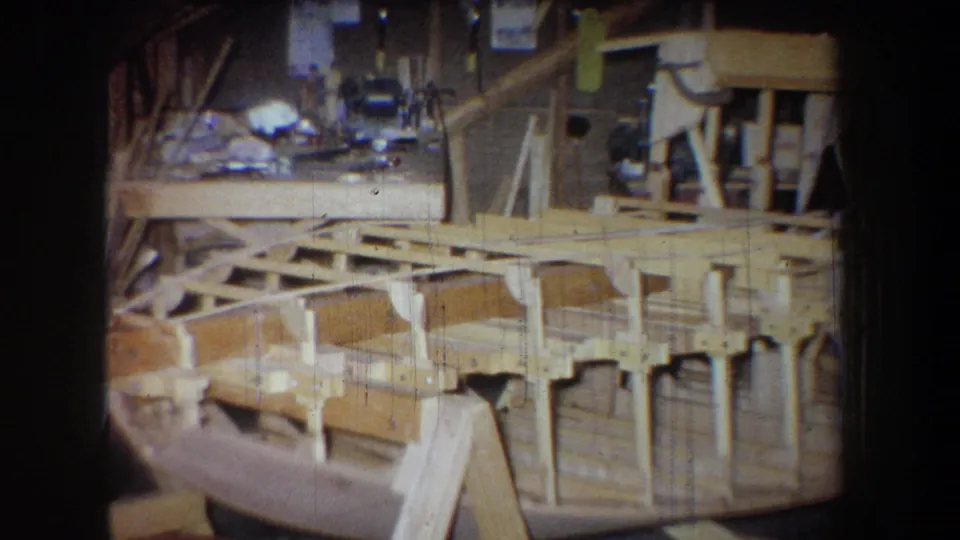 1965 home-movie frame showing the lower curved wooden framework in a private workshop, with vertical supports and tools in the background.