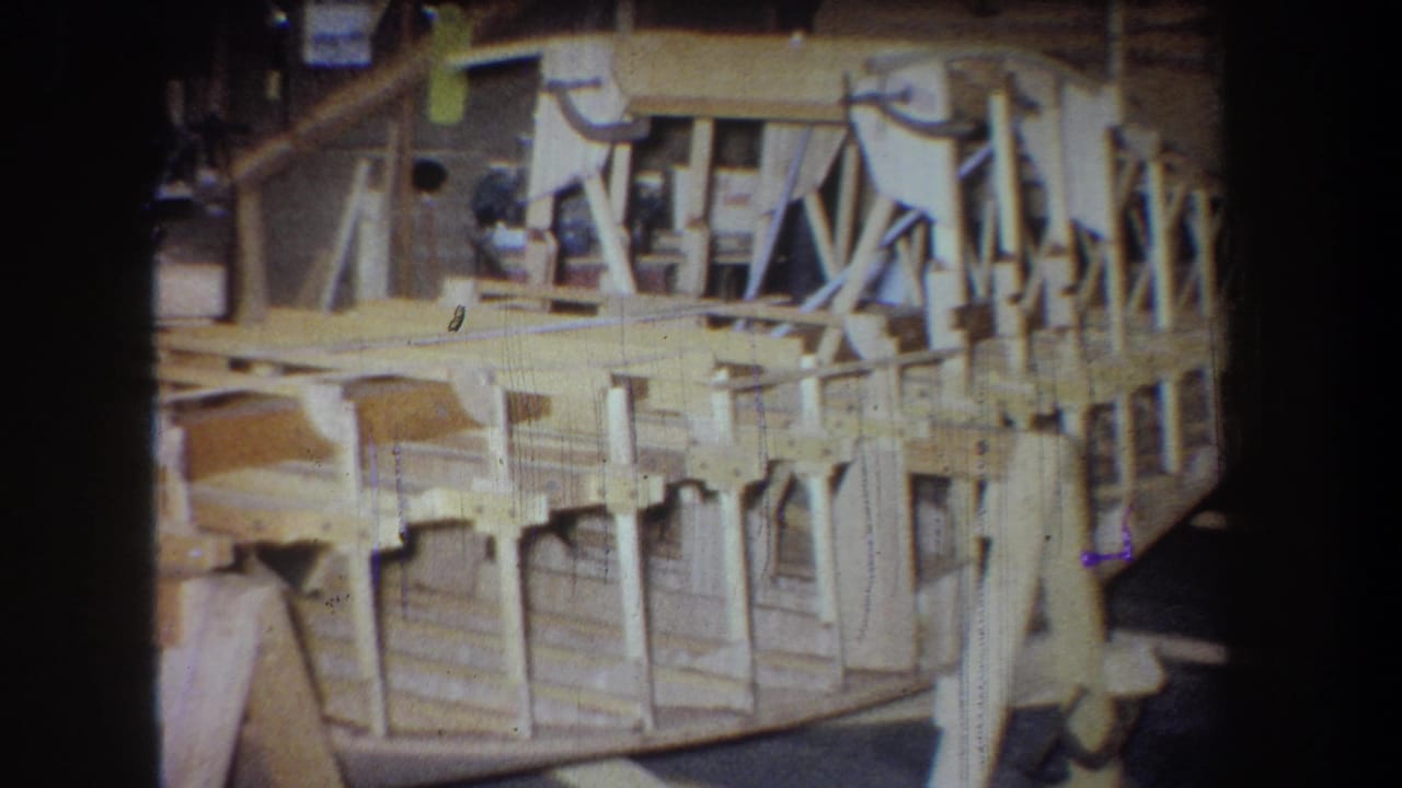 1965 Yucaipa 8mm frame showing a light-colored wooden airplane structure supported inside a workshop.