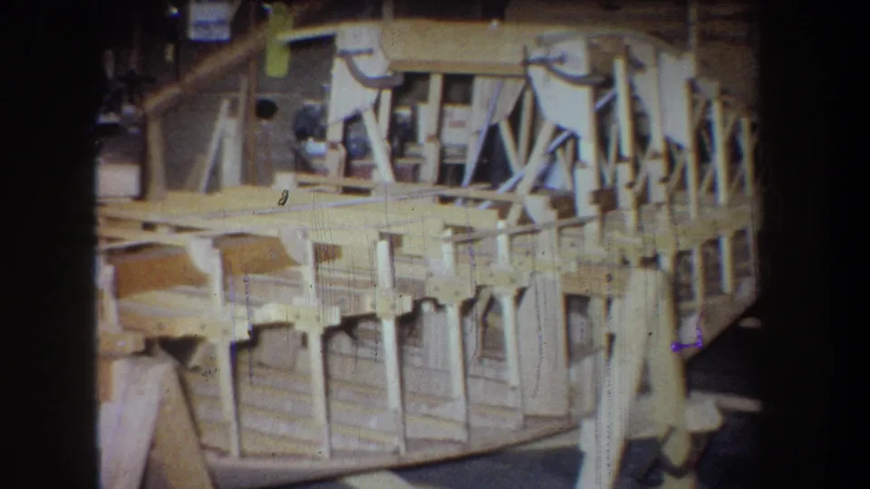 1965 Yucaipa 8mm frame showing a light-colored wooden airplane structure supported inside a workshop.