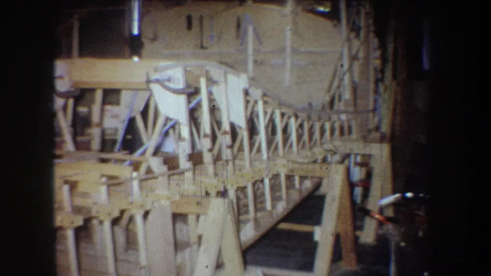 Closer 8mm view of the unfinished wooden airplane frame, showing ribs, braces, and a dark workshop floor opening.