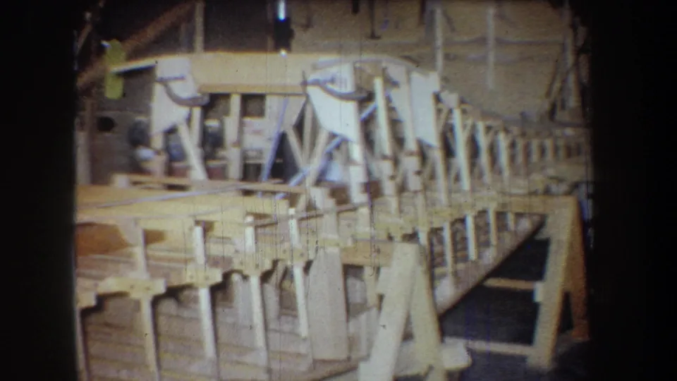1965 8mm frame from Yucaipa showing a wooden boat hull under construction inside a cluttered workshop.