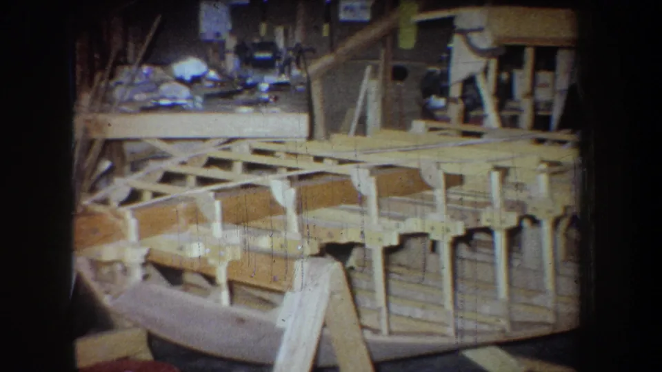 A 1965 8mm workshop view shows the wooden hull frame, exposed supports, and a partially visible adult near the lower edge.