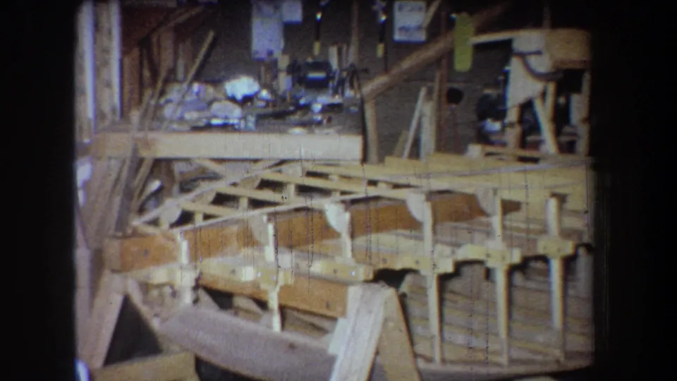 Late in the 1965 Yucaipa clip, the workshop view holds on the braced wooden boat hull with a partial adult figure at left.