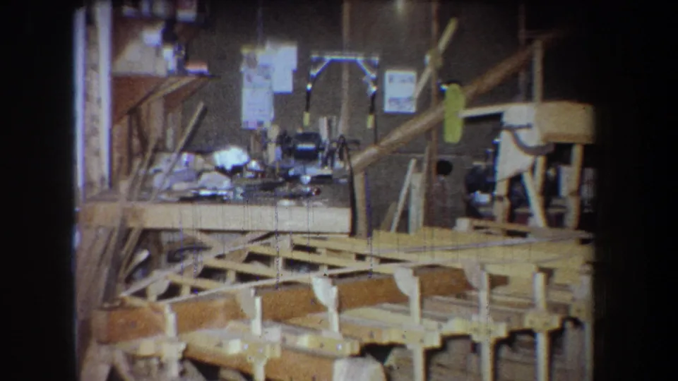 1965 Yucaipa home-movie frame showing a wooden bridge under construction among workbenches, tools, and shop clutter.