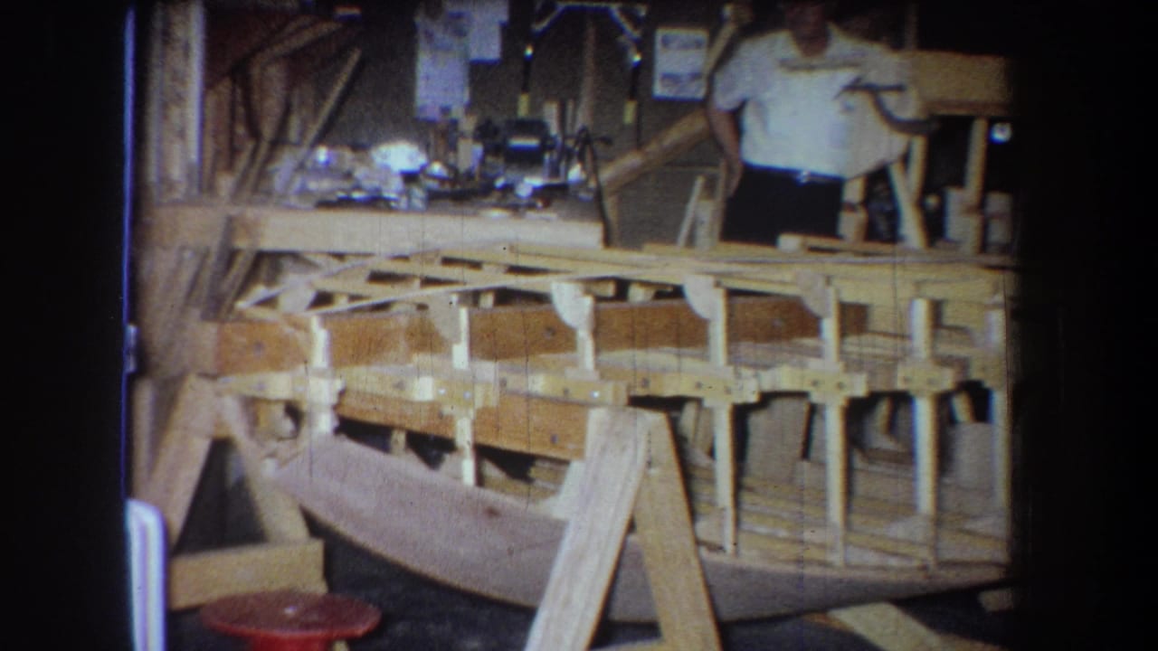 1965 home-movie frame from Yucaipa area showing a man beside a wooden boat hull and support framing in a workshop.