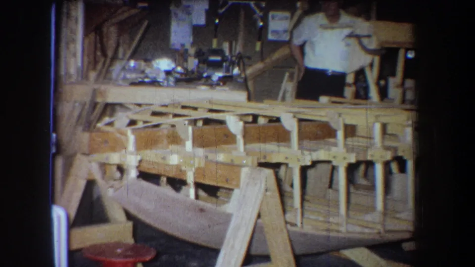 1965 home-movie frame from Yucaipa area showing a man beside a wooden boat hull and support framing in a workshop.