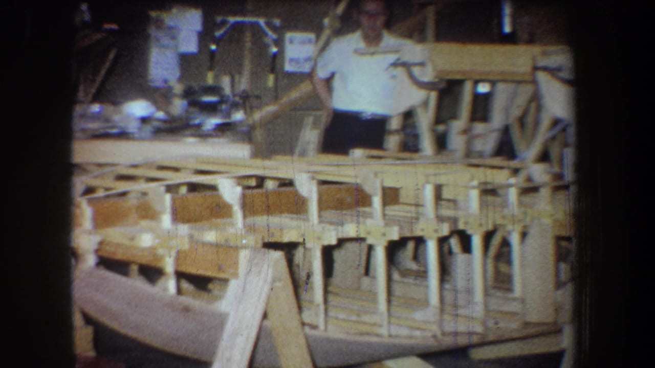 1965 workshop footage shows a man partly obscured by a wooden boat hull, with shop materials and framing behind him.