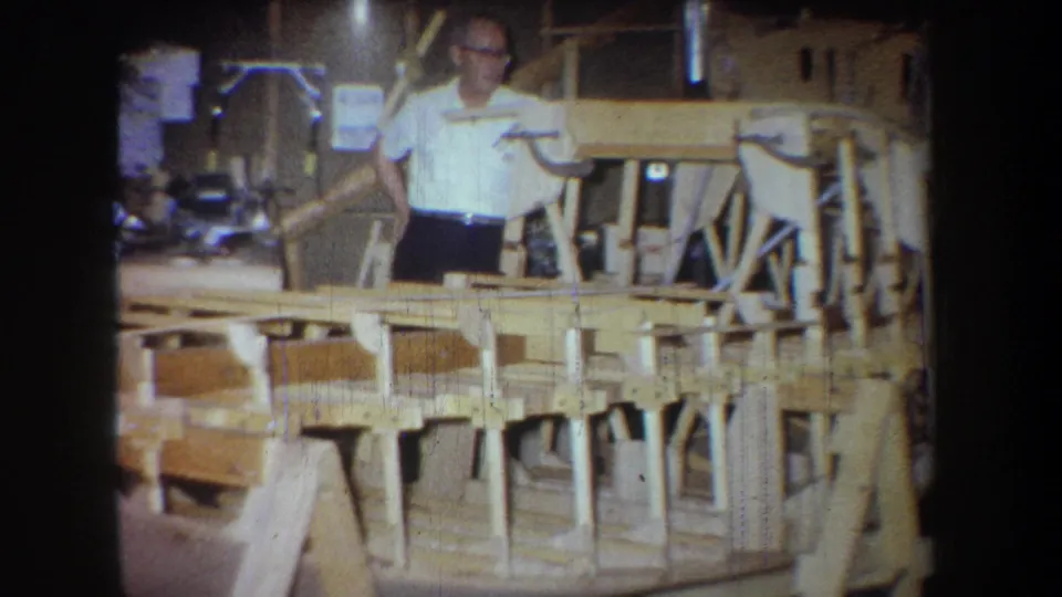 1965 home-movie frame shows a man in a white shirt looking down toward a long wooden boat frame in a private workshop.