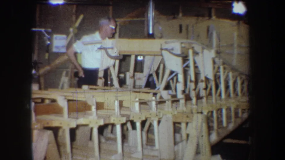 1965 Yucaipa-area home-movie frame shows a man inspecting or standing beside a long wooden boat hull on shop supports.