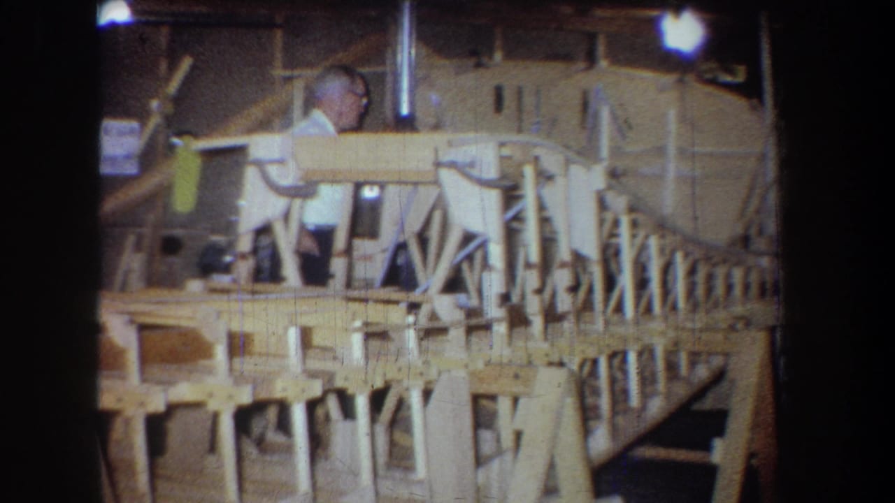 In 1965 Volmer, Yucaipa, California, an older man stands near a large wooden model structure inside a warm-lit workshop or garage.