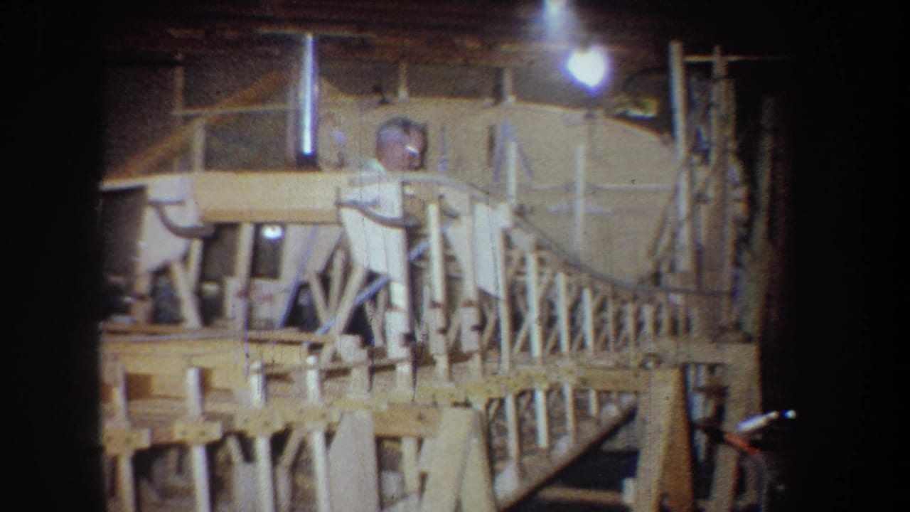 The man appears partly obscured by the wooden model framework, with sawhorses and warm overhead light visible in the 1965 home-movie frame.