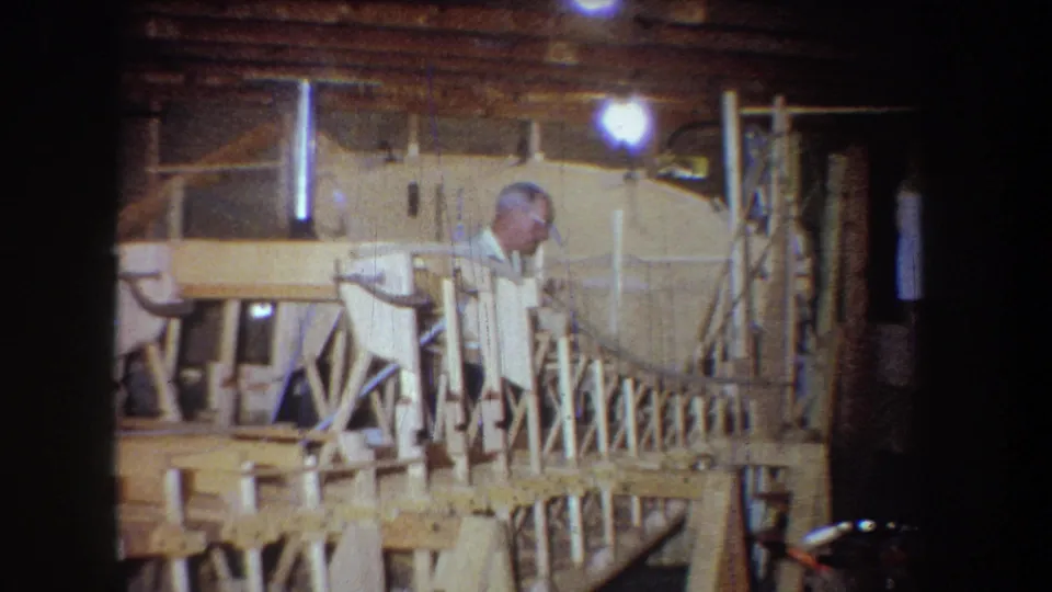 An older man in a white shirt leans near the wooden model structure in a warm-lit private workshop or garage in Volmer, Yucaipa.